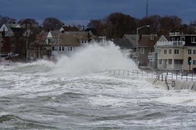 Large waves crash against a seawall in a coastal neighborhood, sending sprays of water high into the air.