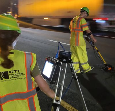 Two workers in high-visibility yellow safety vests and green hard hats conduct a nighttime road survey.