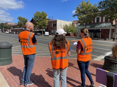 Three individuals are standing on a sidewalk in an urban area, wearing orange safety vests with the logo and text "UConn CLEAR" printed on the back. They appear to be engaged in a field assessment or survey, discussing and possibly taking notes as they observe the surrounding environment.