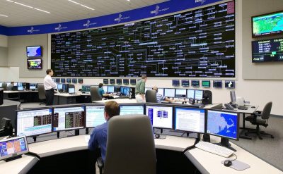 A large control room with several workers seated at desks, each with multiple computer monitors displaying various data and maps.