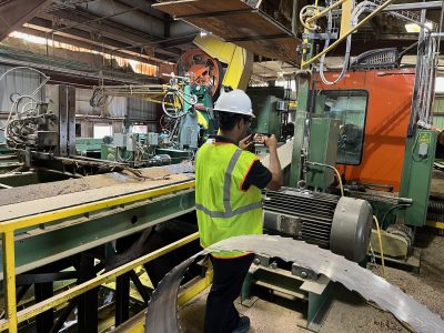 A worker conducting an assessment inside an industrial facility. Wearing a white hard hat and neon safety vest, the worker uses a smartphone to document the operations of large, complex machinery, likely part of a manufacturing or production line.