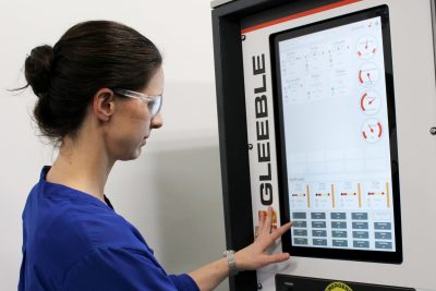 A woman in a blue lab coat and safety glasses operating a sophisticated machine labeled "Gleeble." The woman is intently focused as she interacts with the touchscreen interface of the machine, which displays various controls and real-time data, such as force, temperature, and stress measurements.