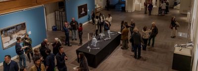 A gathering at a cultural exhibition. The room is filled with people engaged in conversation, observing art, and exploring the displayed works.