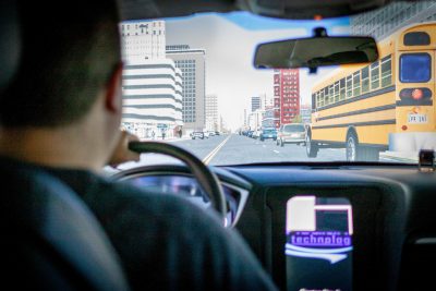 A person seated in a driving simulator, providing a first-person perspective as they navigate through a virtual cityscape. The view through the windshield shows a busy urban street lined with tall buildings, vehicles, and a school bus in the adjacent lane. The simulated environment appears realistic, designed to replicate the experience of driving in a city, with the buildings' detailed facades and the layout of cars creating a sense of immersion.