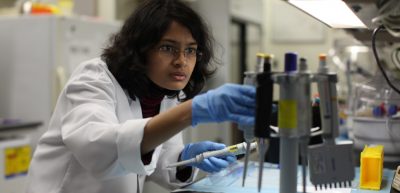 A dedicated researcher in a lab setting, fully focused on her task. She is wearing a white lab coat and blue gloves, handling a pipette with precision.