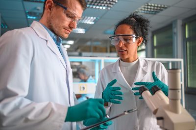 Portrait of mid adult female and male students controlling the test results on a note pad. Both wearing lab coats, latex gloves and protective glasses. Looking away, waist up shot with lab equipment in background, a  microscope in foreground.