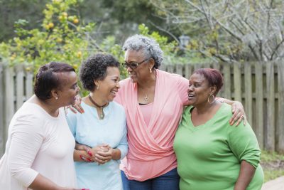 A group of four mature African-American women standing together outdoors in the back yard, side by side, smiling and talking. The tall one is a senior woman in her 60s. Her friends are in their 50s.