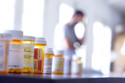 A large group of prescription medication bottles sit on a table as a man in the background stands with his hand on his head. The image is photographed with a very shallow depth of field with the focus being on the pill bottles in the foreground.