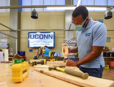 A student wearing a protective mask and safety goggles works on a woodworking project in the UConn Learning Community Innovation Zone.