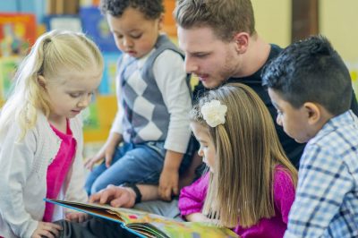 An adult male is reading a colorful book to a group of four young children, who are gathered closely around him.