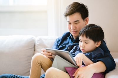 A boy sits on his father's lap as they play on a tablet together