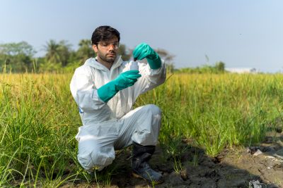 A scientist wearing protective white coveralls, green gloves, and safety glasses is crouched down in a field collecting a water sample into a small container.