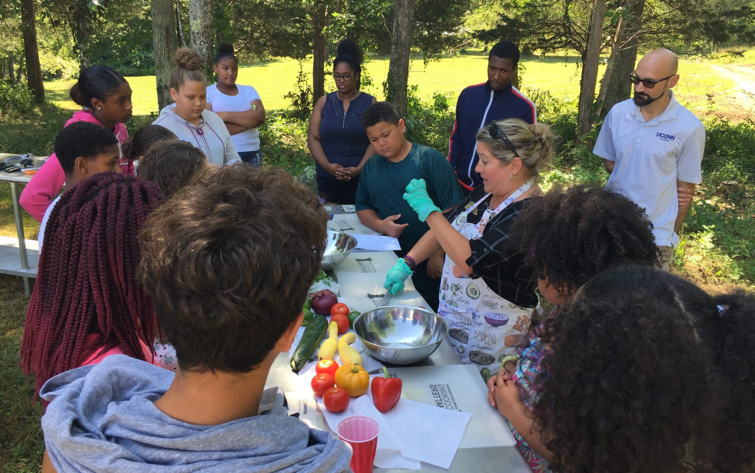 A community partnership event where an instructor leads a hands-on outdoor cooking demonstration for a group of diverse children and teens.