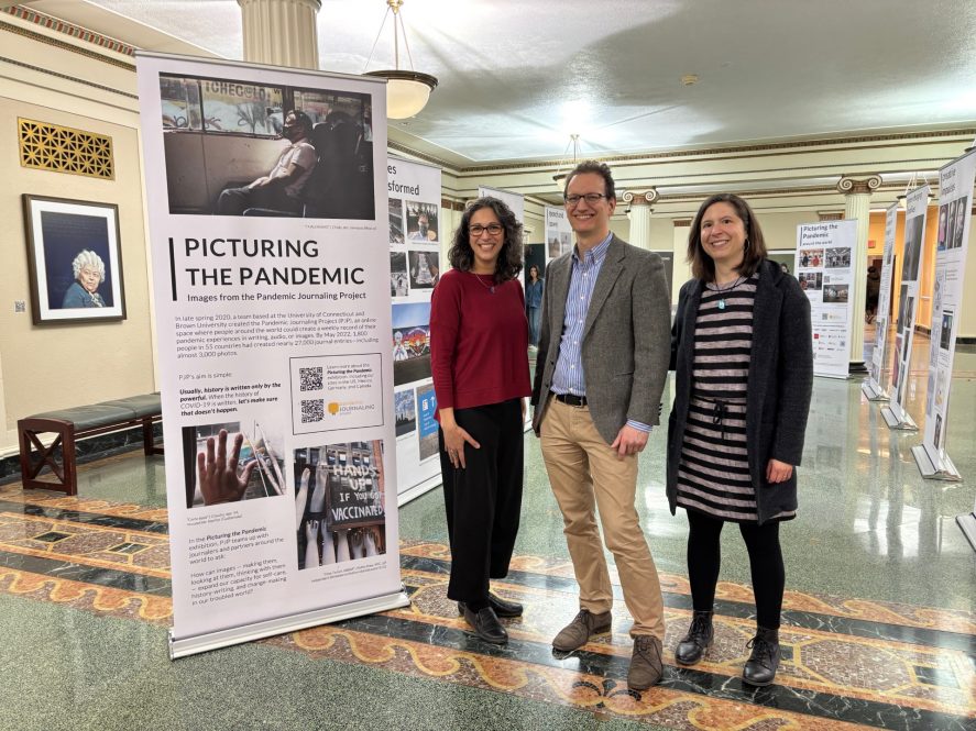 Three people sanding in a gallery of standing posters, one stating "Picturing the Pandemic"