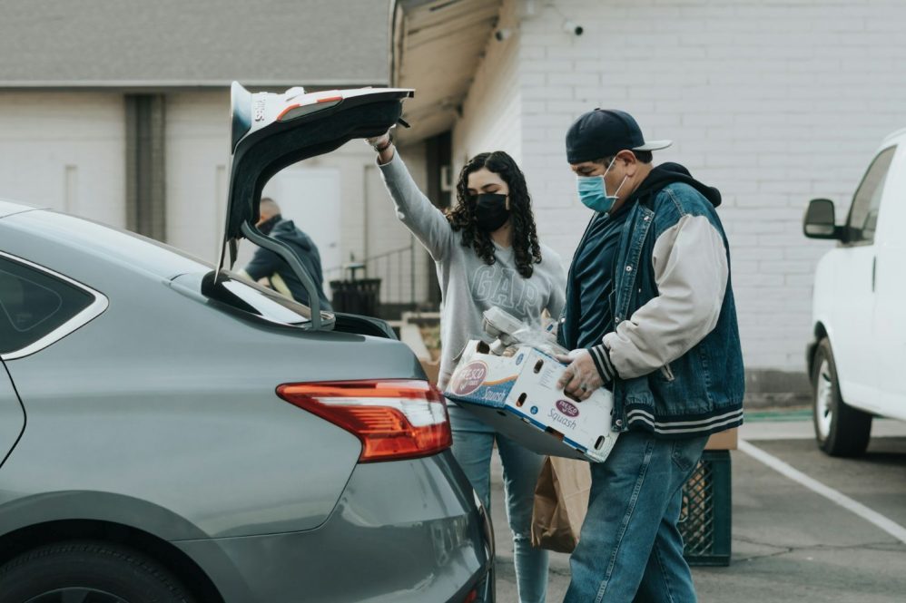 a man and woman wearing health masks load food into a car trunk